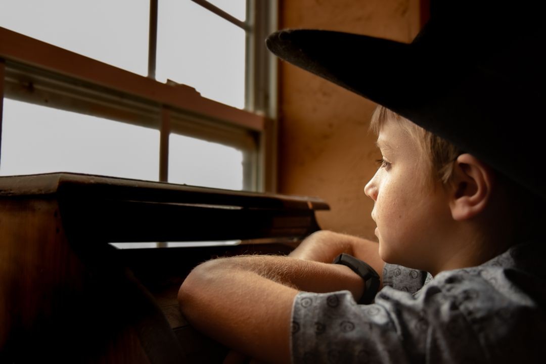 Child wearing a cowboy hat gazing out of a window with thoughtful expression, leaning on arms.